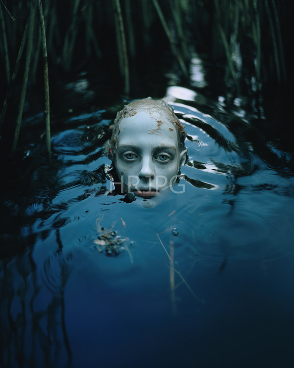 High Resolution: Pale bald androgynous person with strikingly white skin and wide intense blue eyes is partially submerged in dark reflective water surrounded by tall objects download. | HRJPG.com