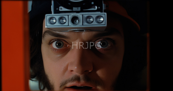 High Resolution: Close-up photograph of actor Jake Gyllenhaal with wide intense blue eyes and dark brown hair wearing an orange cap adorned with vintage camera equipment download. | HRJPG.com