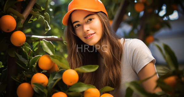 High Resolution: Young woman with long brown hair wearing an orange baseball cap and white t-shirt smiling gently while surrounded by lush green leaves and ripe oranges in a sunlit garden download. | HRJPG.com