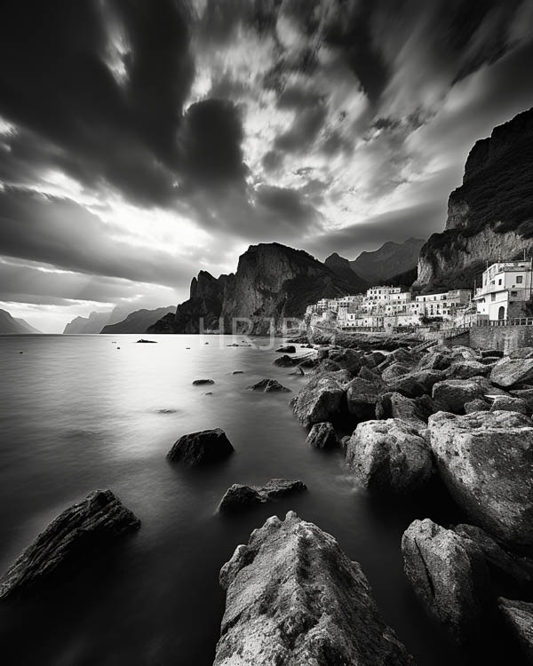 High Resolution: Black-and-white photograph of a rocky coastal landscape at dusk featuring large rocks in the foreground and white-washed buildings on the right with mountains behind download. | HRJPG.com