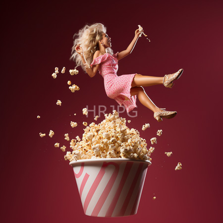 High Resolution: Energetic blonde woman with wavy hair mid-air jumping from above a large striped red-and-white popcorn bucket against a deep maroon background download. | HRJPG.com