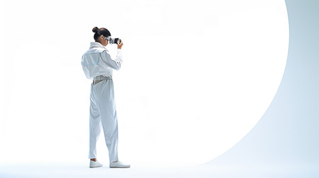 High Resolution: Asian woman dark hair bun white oversized jacket pants set gray sneakers stands side-on holding black digital camcorder download. | HRJPG.com